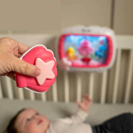 Dłoń trzymająca różowy pilot z gwiazdą, z pozytywką Baby Einstein w tle.